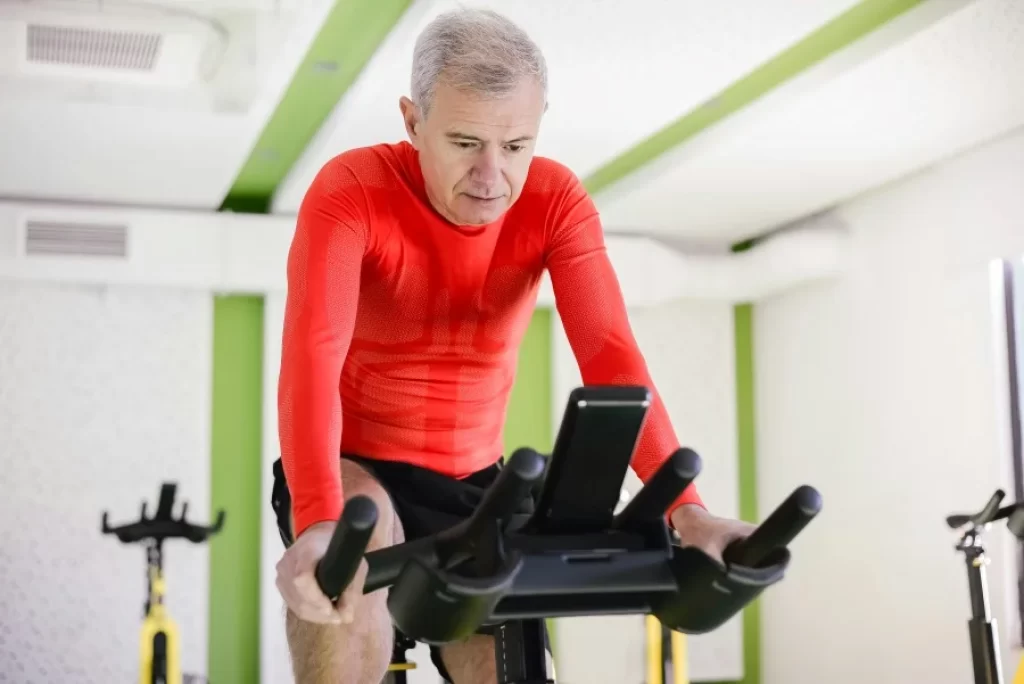 Les bienfaits du vélo d'appartement sont nombreux. Ici, un homme aux cheveux blancs en maillot rouge pédale sur un vélo d'intérieur.